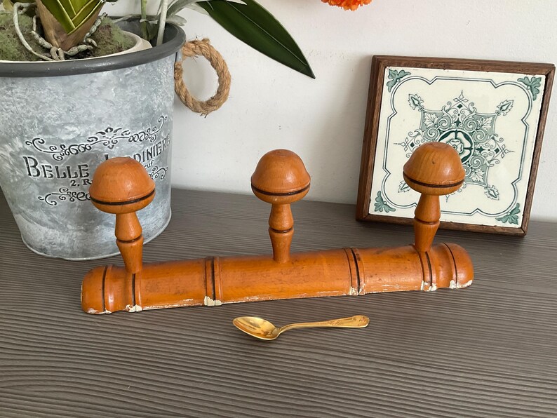 May include: An antique wooden coat rack with three rounded pegs, painted in a warm orange hue. A small gold spoon rests below. A decorative tile and a metal planter with a plant are in the background.