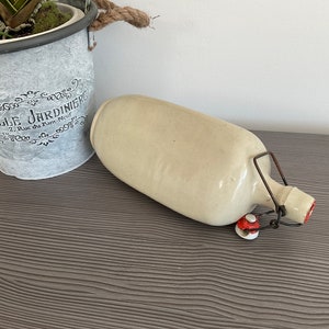 May include: A white ceramic bottle with a metal hinged lid and red accents. The bottle is sitting on a dark wood surface.