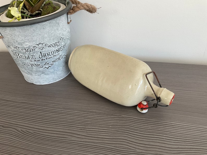 May include: A white ceramic bottle with a metal hinged lid and red accents. The bottle is sitting on a dark wood surface.
