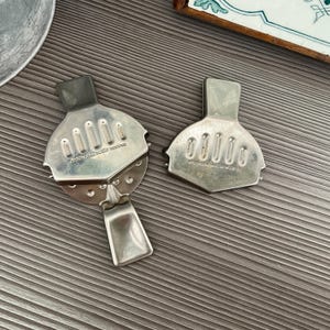 May include: Two vintage silver citrus juicers with a rectangular handle and a semi-circular head with slotted openings. The text "PATER CITRO PATENT ROSTFREI" is visible on the juicers. The juicers are on a wooden surface.