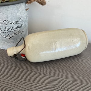 May include: A white ceramic bottle with a metal handle and a red stopper. The bottle is lying on a grey textured surface.