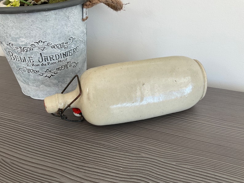 May include: A white ceramic bottle with a metal handle and a red stopper. The bottle is lying on a grey textured surface.