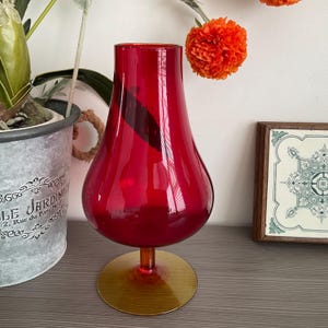 May include: A striking red glass vase with a rounded body and a flared opening. The vase is supported by a circular, amber-toned base. This decorative vase is suitable for displaying flowers or as a standalone decorative item.