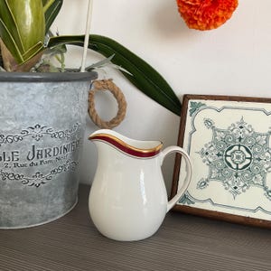 Può includere: Una brocca in ceramica bianca con finiture rosse e dorate. La brocca ha un manico curvo e un beccuccio per versare. La brocca è su una superficie di legno accanto a una piastrella decorativa e un vaso di metallo.