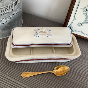 Peut inclure: Un beurrier rectangulaire vintage avec couvercle, crème avec des bordures marron et bleu. Le couvercle est orné d'une poignée et de motifs bleus. Une cuillère dorée est posée devant le plat.