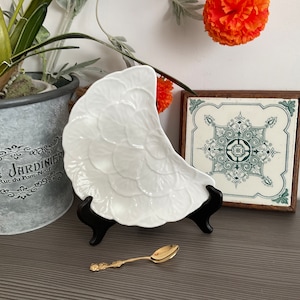 May include: White ceramic half-moon shaped dish with a raised floral design. The dish is on a black stand and a small gold spoon is in front of it.