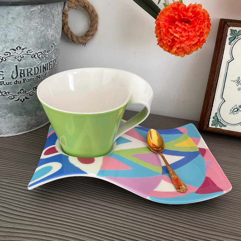 May include: A green and white teacup with a unique handle sits on a colorful, wavy saucer with a gold spoon. The saucer has a geometric pattern in shades of blue, pink, yellow, and green.