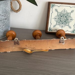 May include: A rustic wooden coat rack with three wooden knobs and metal hooks. A gold spoon rests below the rack. A decorative tile and a metal bucket with the text "LE JARDINIER" are in the background.