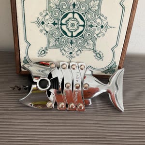 May include: A chrome-finished metal corkscrew shaped like a fish skeleton. The body consists of articulated segments connected by rivets, with a black circular eye and a metal spiral worm. The word LAZY is engraved on one of the central segments.