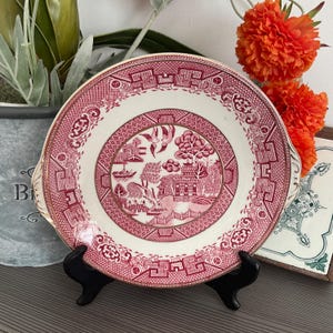 May include: A vintage, round serving plate with a red and white design. The plate features a central landscape scene with a pagoda, trees, and birds, surrounded by a patterned border. It has two small handles and is displayed on a black stand.