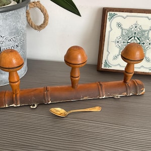 May include: A vintage wooden coat rack with three mushroom-shaped pegs. The rack is a warm brown color with a distressed finish. A small gold spoon rests below the rack. A decorative tile and a metal bucket are in the background.
