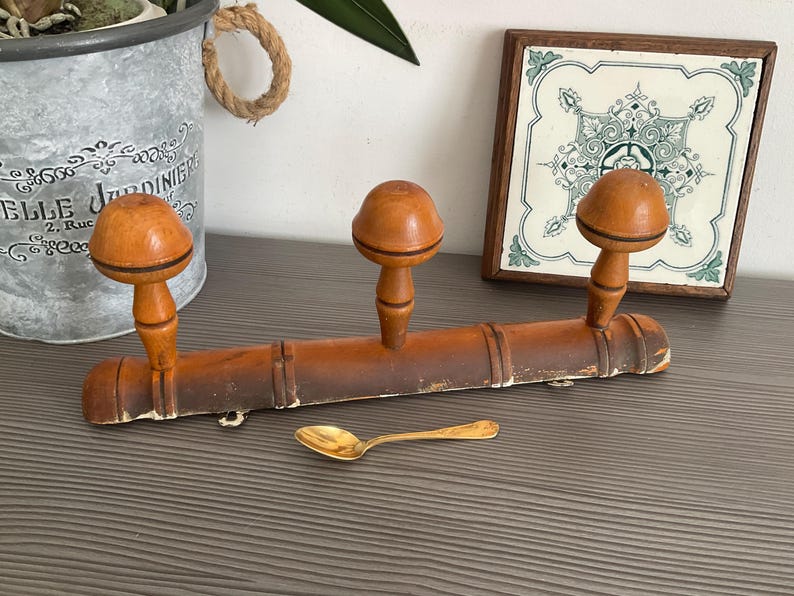 May include: A vintage wooden coat rack with three mushroom-shaped pegs. The rack is a warm brown color with a distressed finish. A small gold spoon rests below the rack. A decorative tile and a metal bucket are in the background.