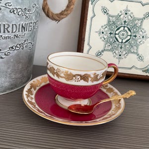 May include: A vintage teacup and saucer set with a gold rim and accents. The cup and saucer are a deep red with gold leaf designs. A small gold spoon rests on the saucer, next to the teacup. The set is on a gray surface.