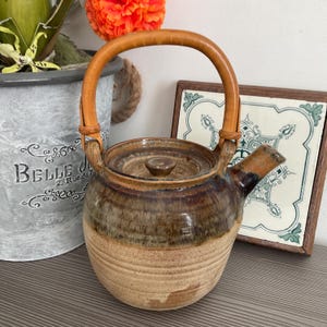 May include: A ceramic teapot with a light brown base and a darker brown upper section. It has a wooden handle and a matching lid. The spout is dark brown and the teapot is sitting on a wooden surface.