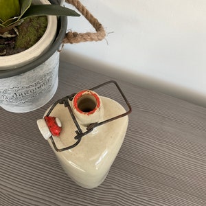 May include: A vintage ceramic bottle with a metal swing top lid and a red and white ceramic stopper. The bottle is a light beige colour and has a rustic, aged look.