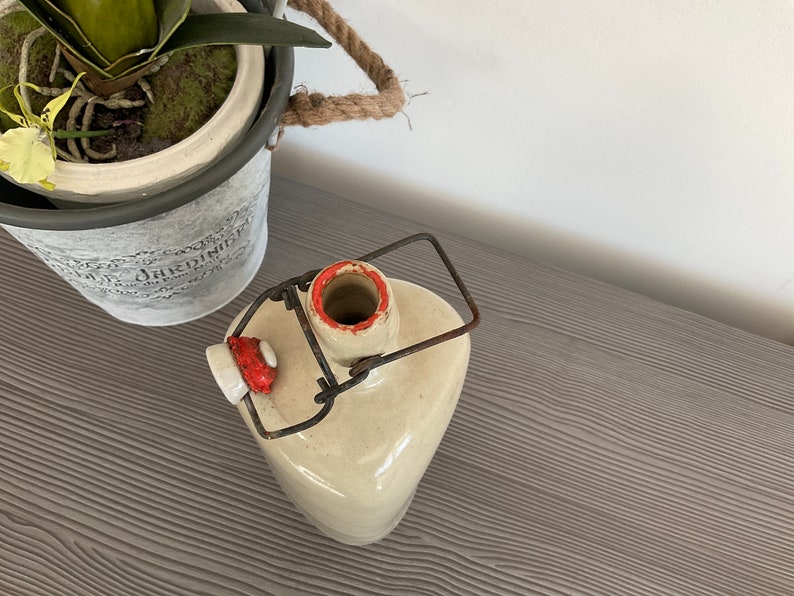 May include: A vintage ceramic bottle with a metal swing top lid and a red and white ceramic stopper. The bottle is a light beige colour and has a rustic, aged look.