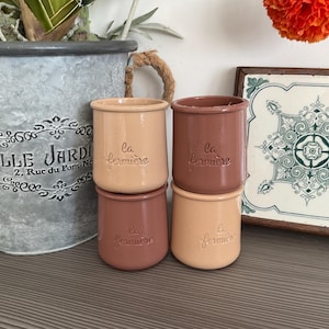 May include: Four small, ceramic pots in shades of beige and brown are stacked. Each pot has the embossed text "La fermière". A galvanized metal planter with the text "LLE JARD" is in the background.