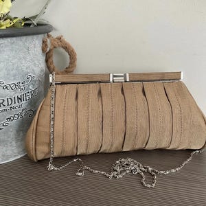 May include: A tan clutch bag featuring a pleated fabric exterior and a silver-toned metal frame closure. The bag includes a detachable, thin silver-toned metal chain strap that rests in front of the pleated body, providing a structured design.