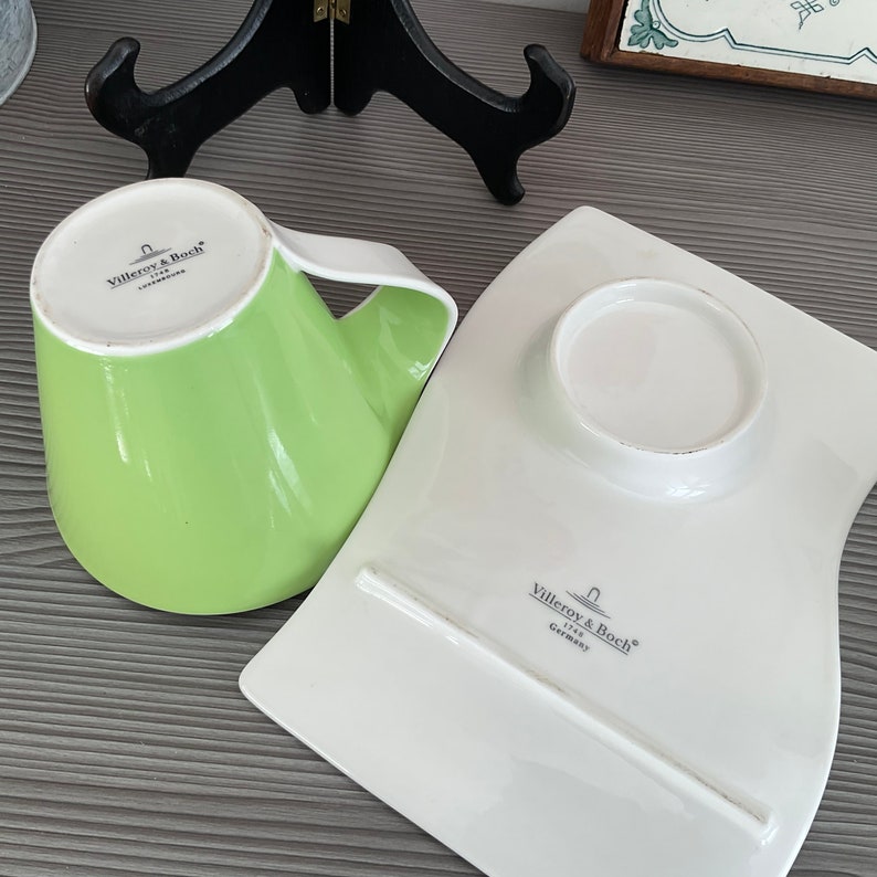 May include: A green and white ceramic teacup and saucer set with the Villeroy & Boch logo. The cup has a unique shape and the saucer is curved.