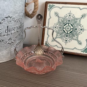 Op de afbeelding: Een roze glazen schaal met een zilveren lepel in een zilveren houder. De schaal heeft een geschulpte rand en een stervormig patroon in het midden. Een decoratieve metalen emmer en een tegel met een bloemenmotief staan op de achtergrond.