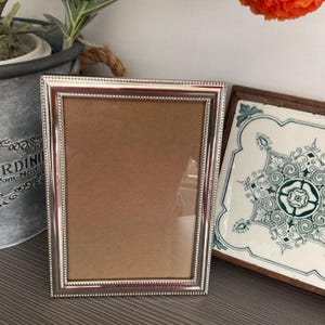 Può includere: Una cornice portafoto rettangolare color argento con un bordo decorato a perline. La cornice presenta una finitura metallica liscia e lucida e un frontale in vetro trasparente, che racchiude un pannello di supporto marrone. Il design è semplice e verticale.