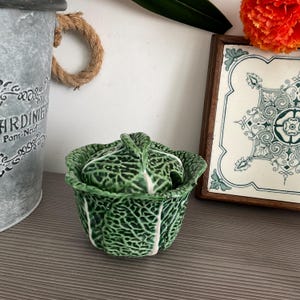May include: A ceramic lidded bowl shaped like a green cabbage head with textured, overlapping leaf details. The bowl features a deep green color with lighter white veins running through the leaves, mimicking the appearance of a real vegetable.