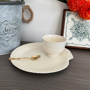 Könnte beinhalten: Ein weißes Tee-Set mit Untertasse und einem goldenen Löffel auf einem weißen, muschelförmigen Teller. Der Teller hat ein geriffeltes Design.