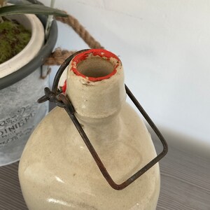 May include: A vintage ceramic bottle with a metal carrying handle. The bottle has a cream-coloured glaze with a red rim around the opening.