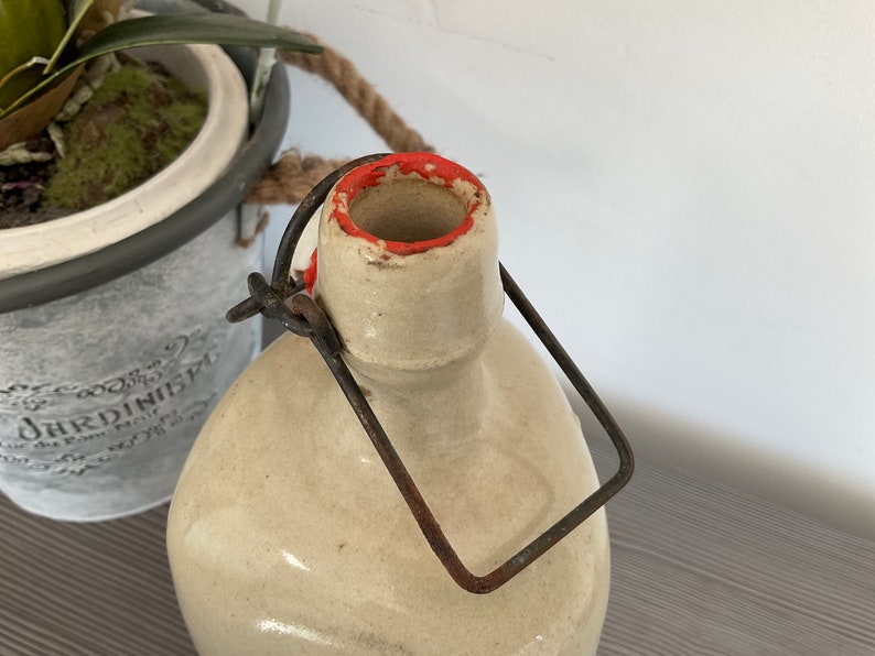 May include: A vintage ceramic bottle with a metal carrying handle. The bottle has a cream-coloured glaze with a red rim around the opening.