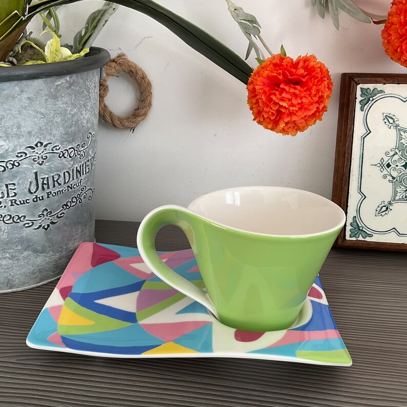 May include: A green and white ceramic teacup with a unique curved handle sits on a colorful geometric patterned saucer.