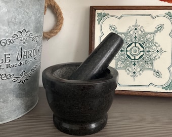 May include: A dark gray stone mortar and pestle set with a smooth, polished exterior and a textured interior bowl. The mortar has a wide, rounded base and a thick rim, while the pestle is cylindrical with a tapered end for grinding ingredients.