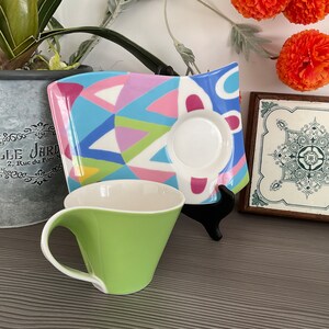 May include: A green and white ceramic teacup with a curved handle and a matching saucer with a colorful geometric design. The saucer has a white center with a blue, pink, and green geometric pattern.