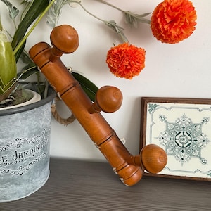 May include: A wooden coat rack with four rounded knobs. The rack is brown and angled, with a decorative plant in a metal pot and a framed tile with a green and white design. Orange flowers are in the background.