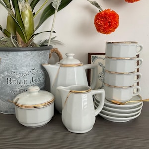 May include: A white porcelain tea set with gold trim, including a teapot, creamer, sugar bowl, and cups. The set is arranged with saucers and spoons, creating an elegant display. The background includes a decorative plant and a framed picture.