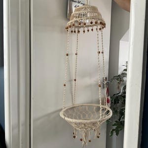 以下が含まれることがあります： クリーム色のマクラメハンギングプランター。段になった上部と織りバスケットのベースが特徴です。木製のビーズが吊り下げコードと上部の周りに散りばめられています。小さな植物を入れるように設計されています。