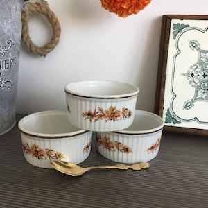 May include: Three white ceramic ramekins with a floral design in shades of orange and brown. The ramekins are arranged in a triangular shape on a grey surface. A gold spoon is in the foreground.