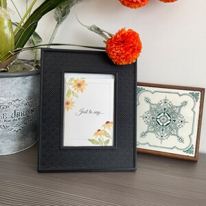 May include: A black picture frame with a white mount and a watercolour print of yellow flowers. The print says "Just to say..." The frame is sitting on a wooden surface with a decorative tile and a potted plant.
