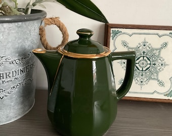 May include: A dark green ceramic teapot with a gold rim and handle. The teapot has a geometric shape and a lid with a gold knob. The spout is angled, and the handle is square. The teapot is on a wooden surface.