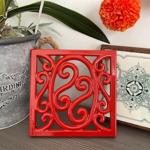 May include: A square, glossy red trivet with an ornate, swirling design. The trivet has a rope loop for hanging. A decorative pot and a framed tile with a floral pattern are in the background.