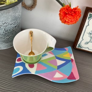 May include: A white teacup with a green base and a gold spoon sits on a colorful, abstract patterned saucer. The saucer has a wavy edge and is decorated with blue, pink, green, and yellow geometric shapes.