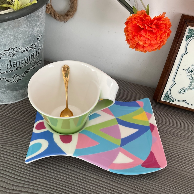 May include: A white teacup with a green base and a gold spoon sits on a colorful, abstract patterned saucer. The saucer has a wavy edge and is decorated with blue, pink, green, and yellow geometric shapes.