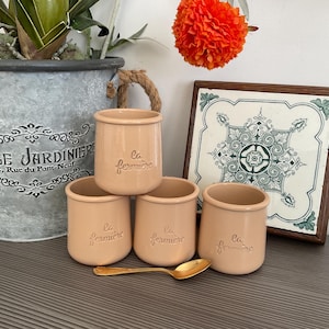 Puede incluir: Cuatro tarros de cerámica color melocotón con el texto "La fermière" en relieve en la parte delantera. Una cuchara dorada descansa frente a los tarros. Una baldosa decorativa y una maceta de metal están en el fondo.