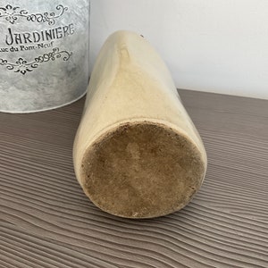 May include: A vintage, off-white ceramic crock with a wide opening and a slightly rounded bottom. The crock has a rough, textured surface and is sitting on a wooden surface.