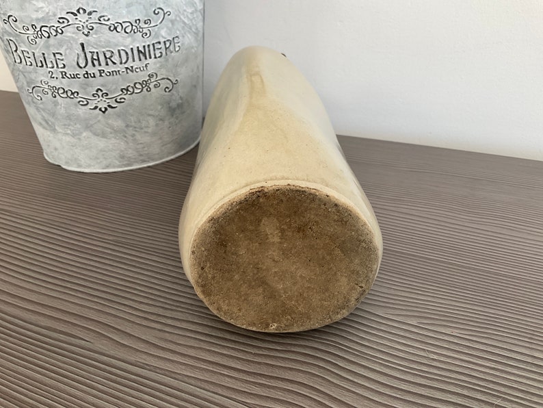 May include: A vintage, off-white ceramic crock with a wide opening and a slightly rounded bottom. The crock has a rough, textured surface and is sitting on a wooden surface.