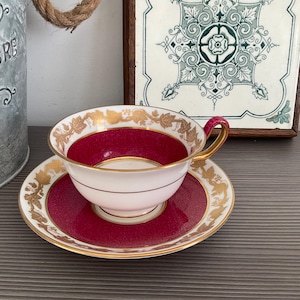 Puede incluir: Un juego de taza y platillo de té vintage. La taza y el platillo tienen una base blanca con un interior y borde rojo intenso. Los detalles de hoja de oro adornan el borde y los bordes del platillo. El asa es de color rosa intenso.