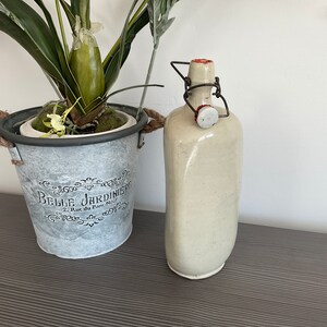 May include: A white ceramic bottle with a metal wire closure and a red rubber stopper. The bottle is sitting on a dark grey surface next to a metal planter with the text "Belle Jardiniere" printed on the side.