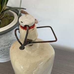 May include: A vintage ceramic bottle with a white lid and a rusty metal handle. The bottle is beige with a light brown glaze.