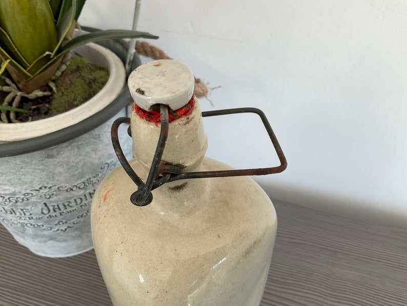 May include: A vintage ceramic bottle with a white lid and a rusty metal handle. The bottle is beige with a light brown glaze.