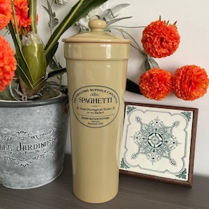 Peut inclure: Un grand pot à spaghetti crème avec un couvercle. Le pot porte une étiquette indiquant "The Original Suffolk Canister Spaghetti" et "Henry Watson Pottery Founded 1800". Le pot est à côté d'une tuile décorative et de fleurs orange.