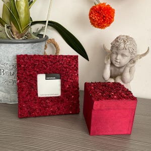 Könnte beinhalten: Ein roter, mit Pailletten besetzter Fotorahmen und eine passende quadratische Box. Der Rahmen hat einen weißen Platz für ein Foto, das ca. 7,5 x 7,5 cm (3x3") groß ist. Im Hintergrund befindet sich eine kleine Engelsstatue.